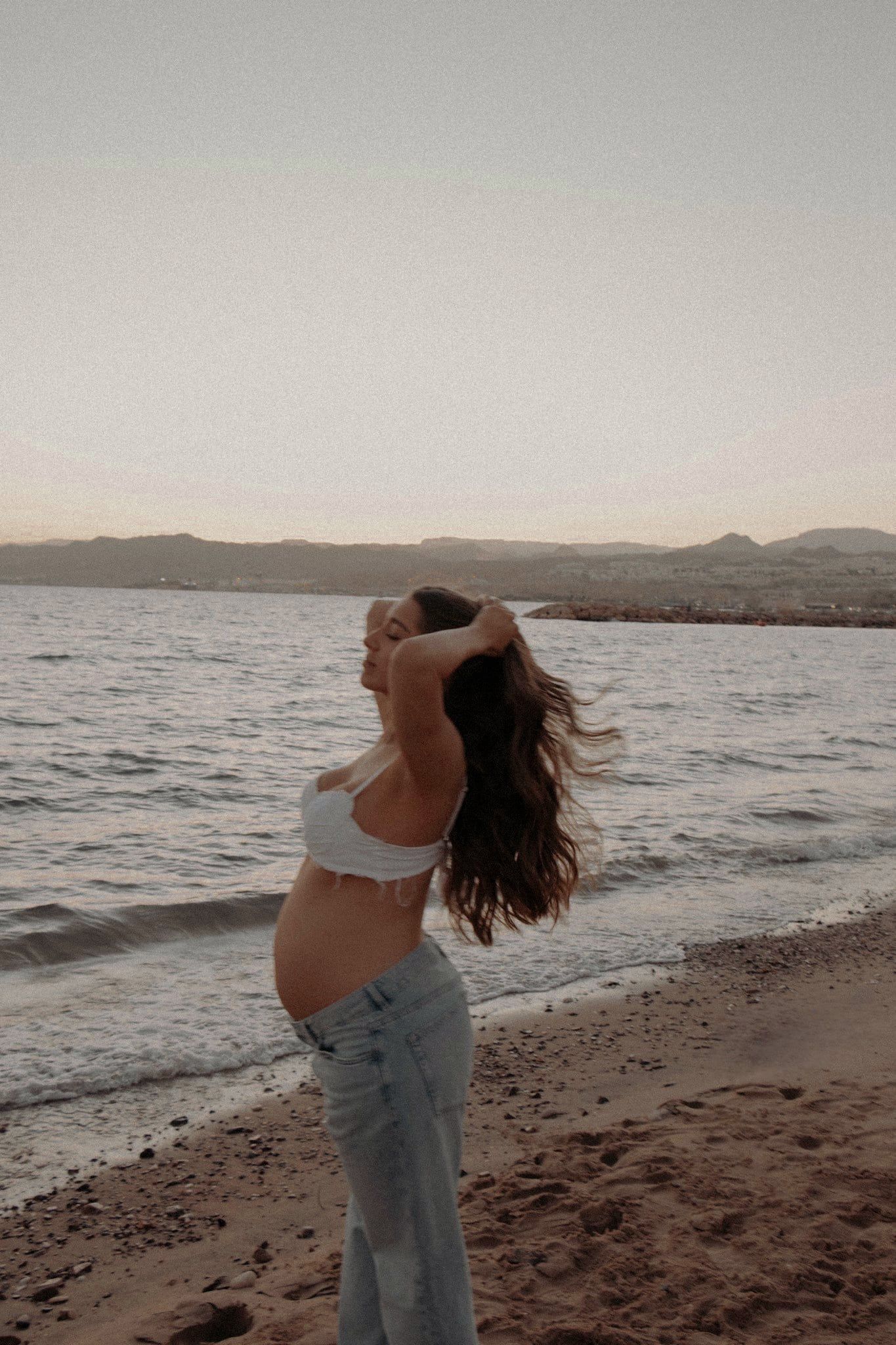 אישה בהריון בג'ינס על החוף באילת, מול המים וההרים